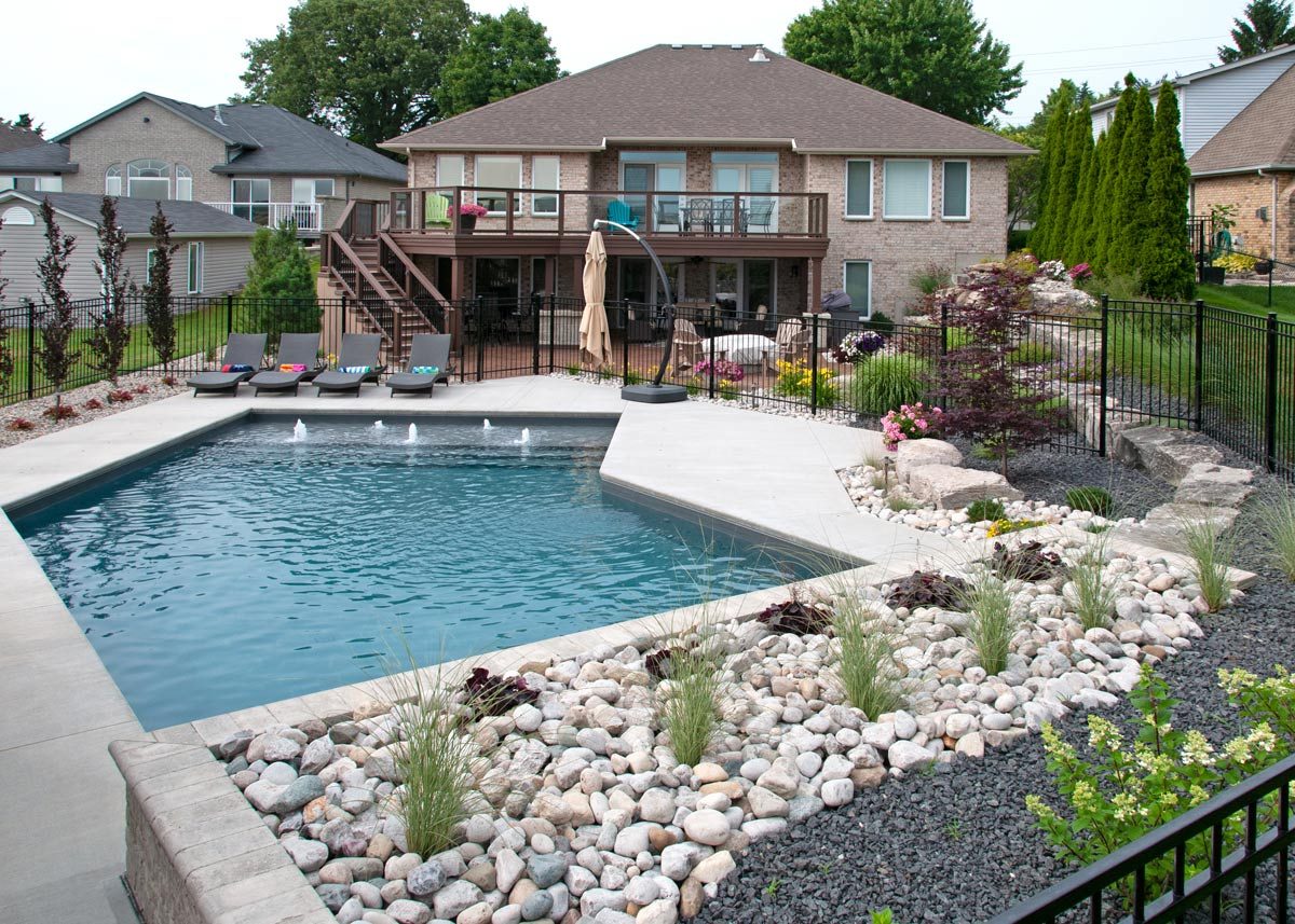Large rectangular inground pool with water features, surrounded by a concrete patio, a rock garden with ornamental grasses, and a black aluminum fence in a residential backyard.