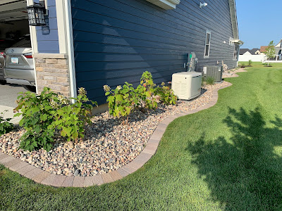 Landscaping bed with river rock and a paver border running alongside a house with blue siding, featuring small green shrubs and an air conditioning unit.