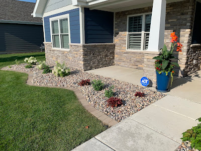 Front yard landscaping with a curved river rock bed, paver border, and small shrubs leading up to a house with blue siding and stone veneer, featuring a blue planter pot on the porch.