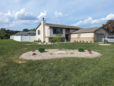 Front yard landscaping featuring a kidney-shaped island bed with river rock, small shrubs, and a central tree, in front of a split-level home with a white privacy fence.