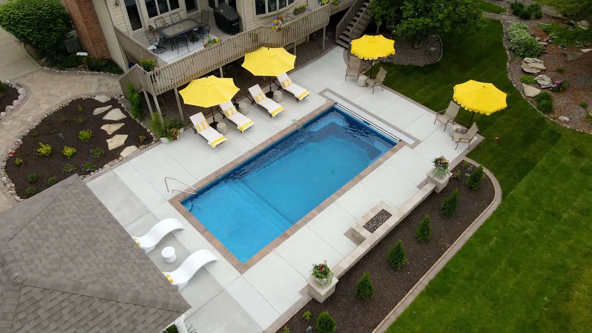 Aerial view of a rectangular inground pool with white concrete deck, yellow market umbrellas, chaise loungers, tanning ledge, automatic pool cover, and curved mulch garden beds by Dean's Pools and Landscaping in Crown Point Indiana