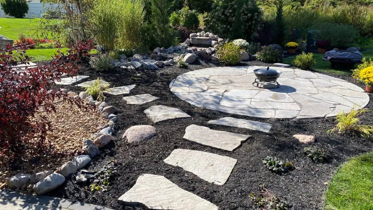 Natural flagstone stepping stone pathway leading to a circular stone patio with a fire bowl, surrounded by lush garden beds, ornamental grasses, and boulders by Dean's Pools and Landscaping