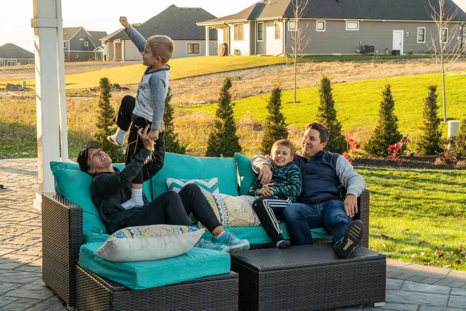 Happy family relaxing and playing on a wicker outdoor sectional sofa on a stone paver patio surrounded by professional landscaping by Dean's Pools and Landscaping in Northwest Indiana