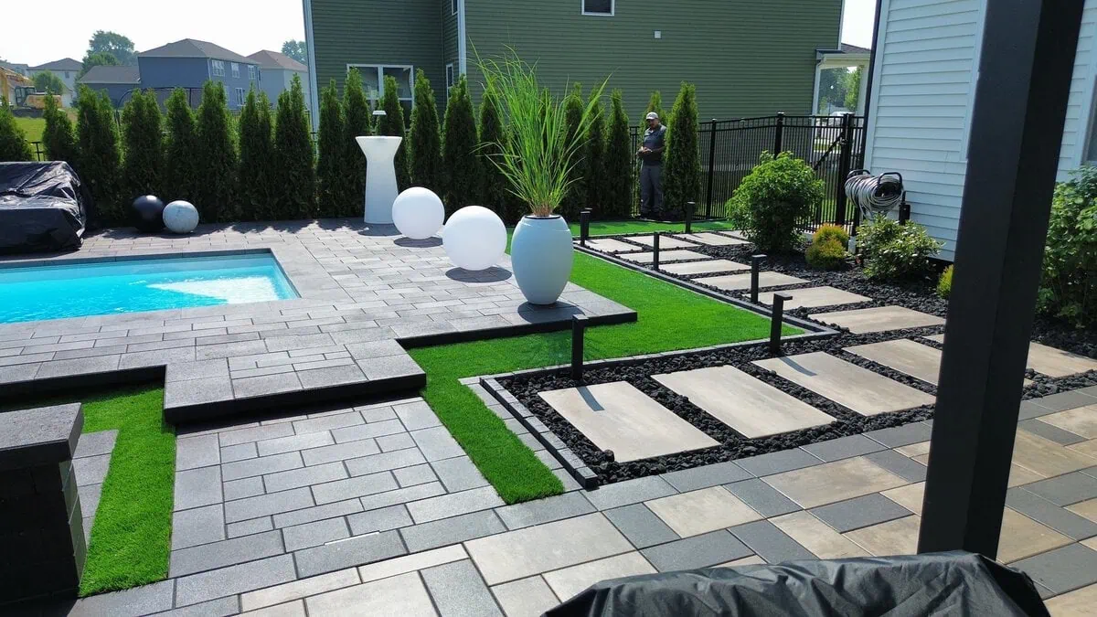 Modern backyard hardscape design with large format concrete pavers, black steel edging, artificial turf, black lava rock stepping stone path, columnar arborvitae privacy hedge, and an inground pool by Dean's Pools and Landscaping