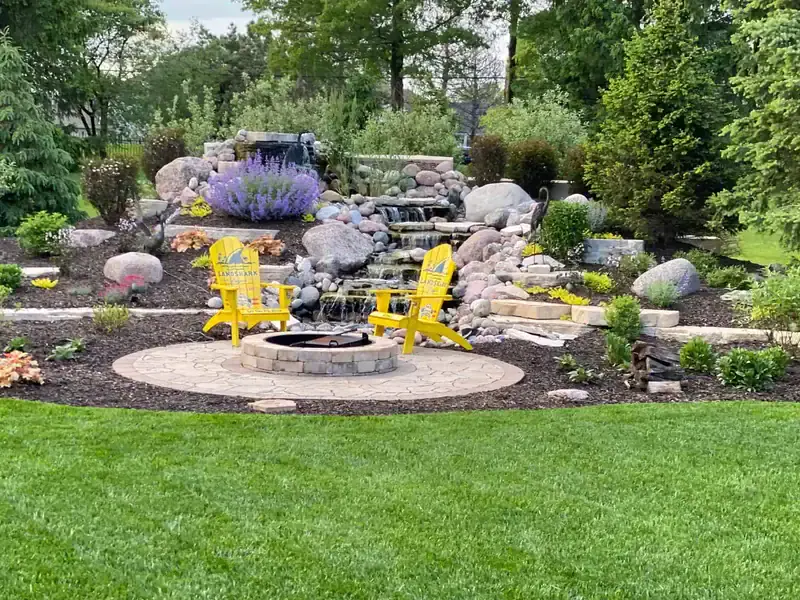 Lush backyard outdoor living space with a round paver fire pit, yellow Adirondack chairs, natural boulder waterfall water feature, colorful perennial garden beds, and manicured lawn by Dean's Pools and Landscaping