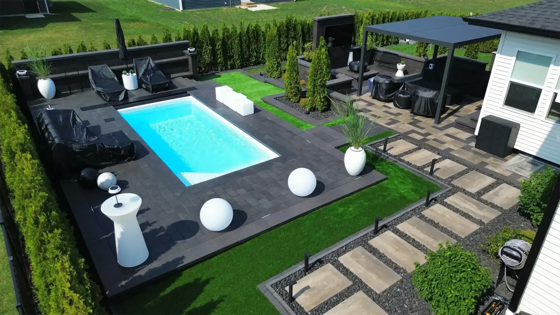Aerial view of a modern luxury backyard with a rectangular inground pool, dark charcoal paver deck, artificial turf, columnar arborvitae, black aluminum pergola, and outdoor kitchen by Dean's Pools and Landscaping