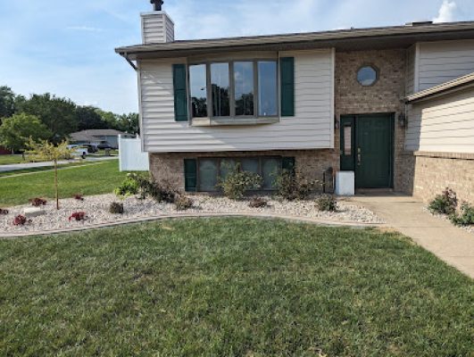 Front yard landscaping showing a curved bed with river rock and small plants leading up to the front entrance of a split-level home with a bay window.