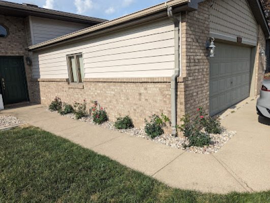 Narrow landscaping bed with river rock and small green plants running alongside a concrete walkway and the brick exterior of a garage.