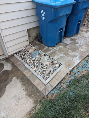Small rectangular landscaping feature filled with decorative river rock and larger stones, bordered by pavers, located next to a concrete walkway and blue trash bins.