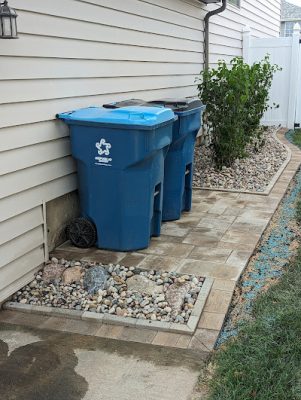 Paver walkway leading to blue trash bins next to a house, with a small rectangular river rock feature and a curved landscaping bed with shrubs.