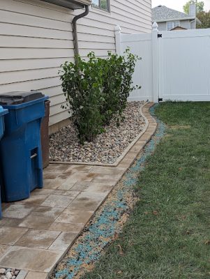Paver walkway alongside a house leading to a white privacy fence, bordered by a curved landscaping bed with river rock and green shrubs.