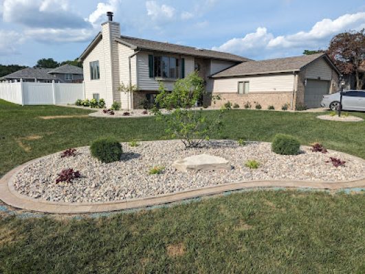 Another view of the front yard landscaping featuring a kidney-shaped island bed with river rock, small shrubs, and a central tree, in front of a split-level home.