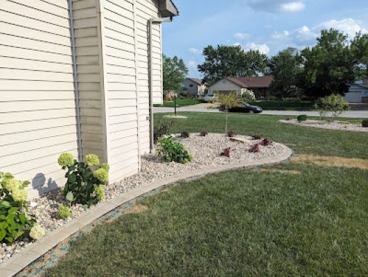 Landscaping bed along the side of a house with light siding, featuring river rock, a paver border, and small green hydrangea bushes.