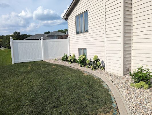 Curved landscaping bed with river rock and a paver border running along the side of a house, featuring small green hydrangea bushes and leading to a white privacy fence.