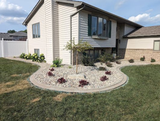 Front yard landscaping featuring a curved bed with river rock, a paver border, small red plants, and a young tree in front of a split-level home.