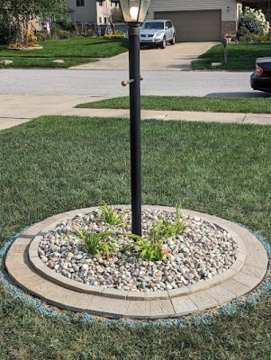 Circular landscaping bed around a black lamppost, filled with decorative river rock, a paver stone border, and small green plants in a front yard.