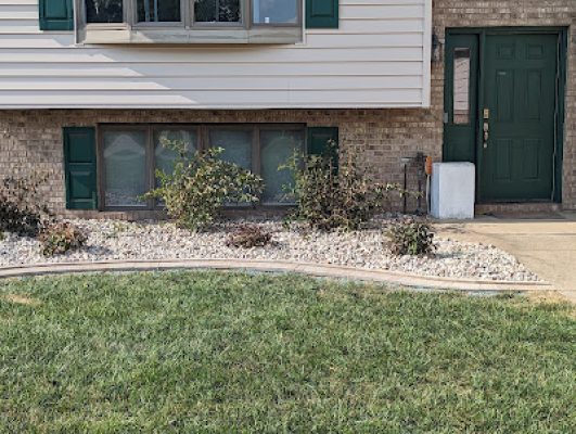 Close-up of a front foundation landscaping bed with river rock, a paver border, and small shrubs planted in front of the lower windows of a split-level home.